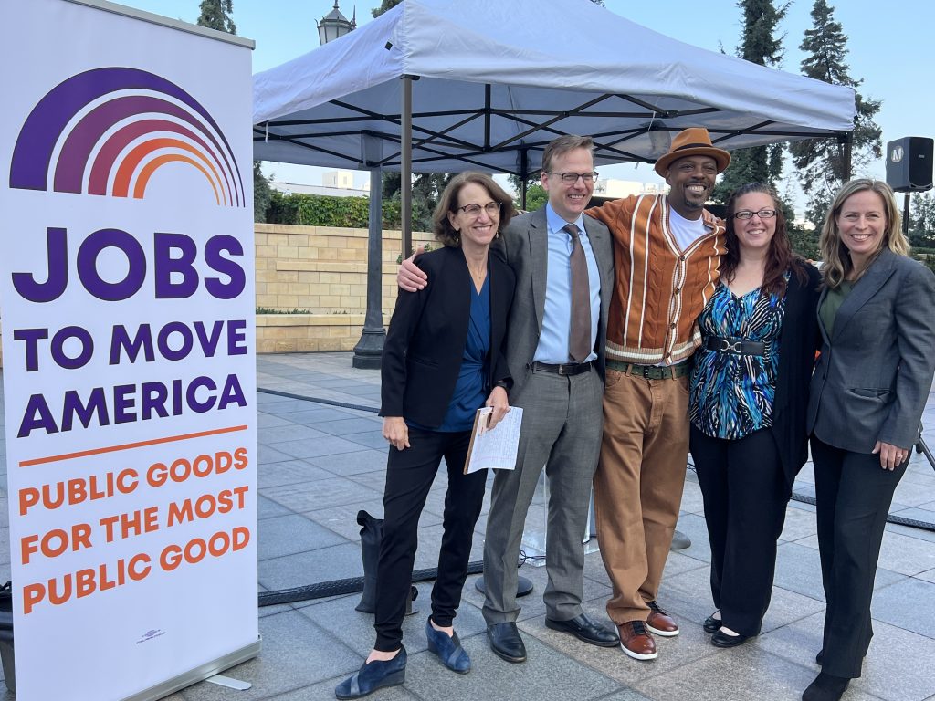 A group of five people stands together, smiling outdoors beneath a tent in front of a banner that reads "Jobs to Move America: Public Goods for the Most Public Good."