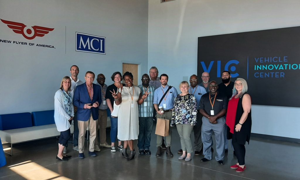 A diverse group of people poses for a photo inside a building, showcasing signs for New Flyer of America, MCI, and the Vehicle Innovation Center. The group stands smiling, with some making peace signs.