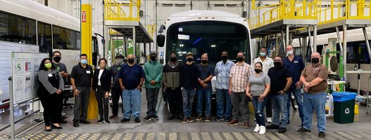 A group of people wearing masks stands in front of a large vehicle inside an industrial facility. They are surrounded by machinery and railings, suggesting a manufacturing environment.